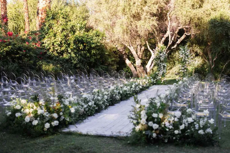   ויקטוריה ואדריאן's outdoor ceremony with ghost chairs, an aisle lined with greenery and white flowers, and a greenery and floral arch