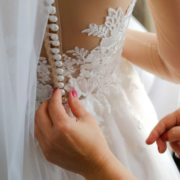   Une demoiselle d'honneur portant du vernis à ongles rose aide la mariée à boutonner sa robe de mariée blanche.