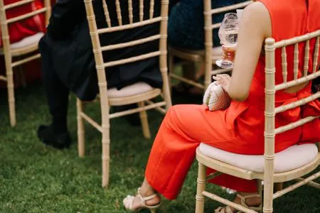   Punahihaista housupukua pukeutunut nainen istuu puisella tuolilla hääseremonian aikana.