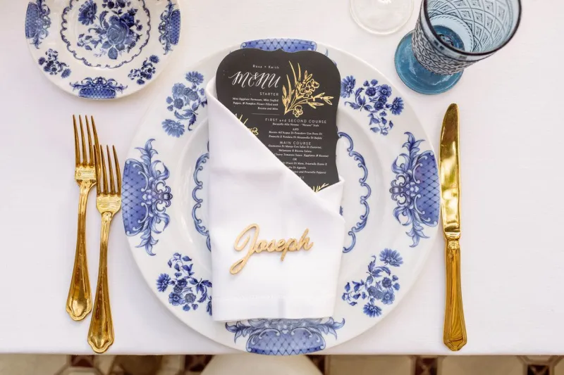   Rosa ja Keith's places set with gold flatware, blue and white chargers, blue goblets, and gold place cards