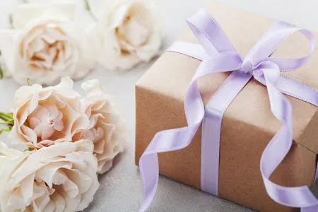   Ruskeaan paperiin kääritty lahja, jossa on violetti rusetti vaaleanpunaisten kukkien vieressä.