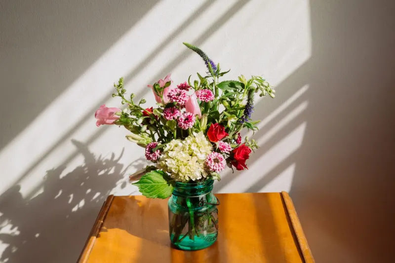   Cvjetni aranžman od crvenih ruža, ružičastog cvijeća i zelenila u plavoj vazi koja stoji unutra na stolu.
